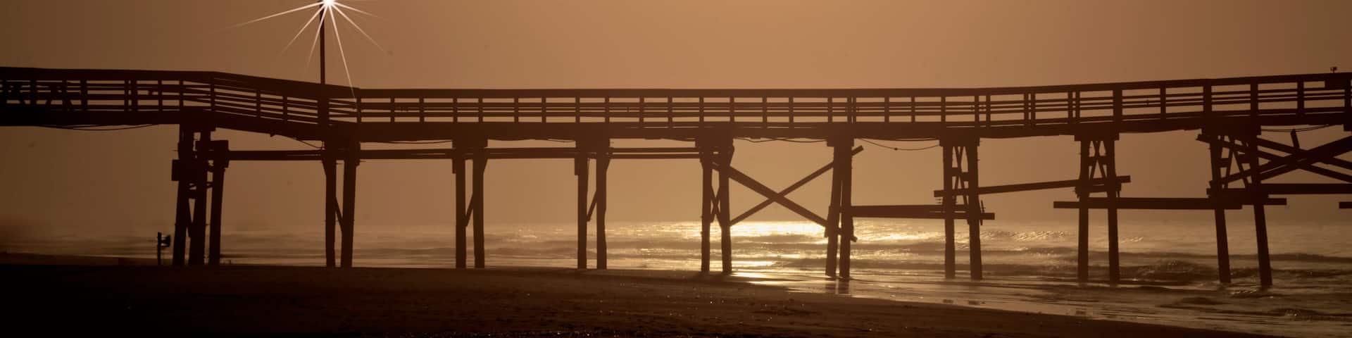 Cherry Grove Fishing Pier The image shows a serene sunrise scene at North Myrtle Beach, South Carolina. A wooden pier stretches across the frame, extending out over the calm ocean waters.