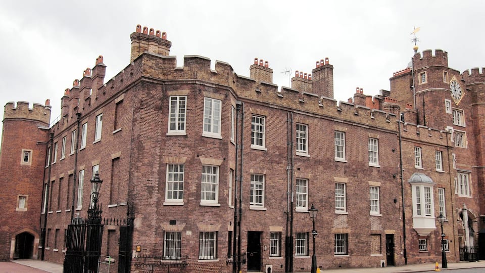 A historic brick building with multiple chimneys, a clock tower, and a gated entrance.