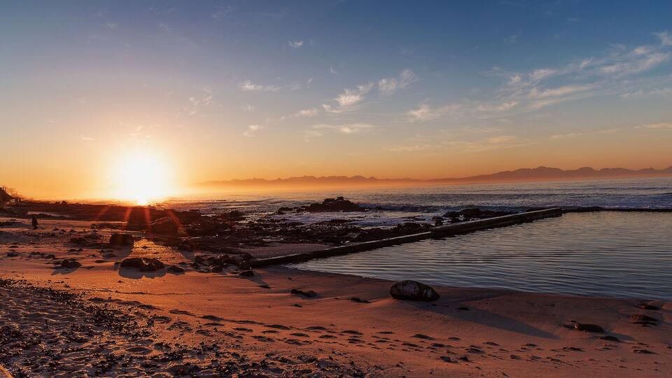 A beach at sunset with the sun low on the horizon, rocky shoreline, and calm water.