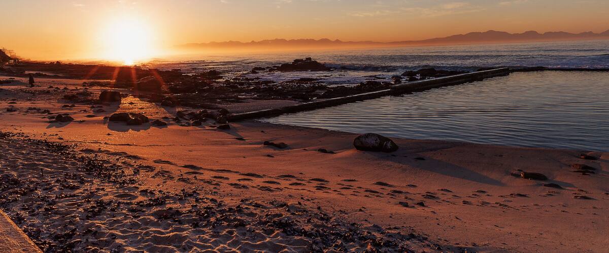 A beach at sunset with the sun low on the horizon, rocky shoreline, and calm water.