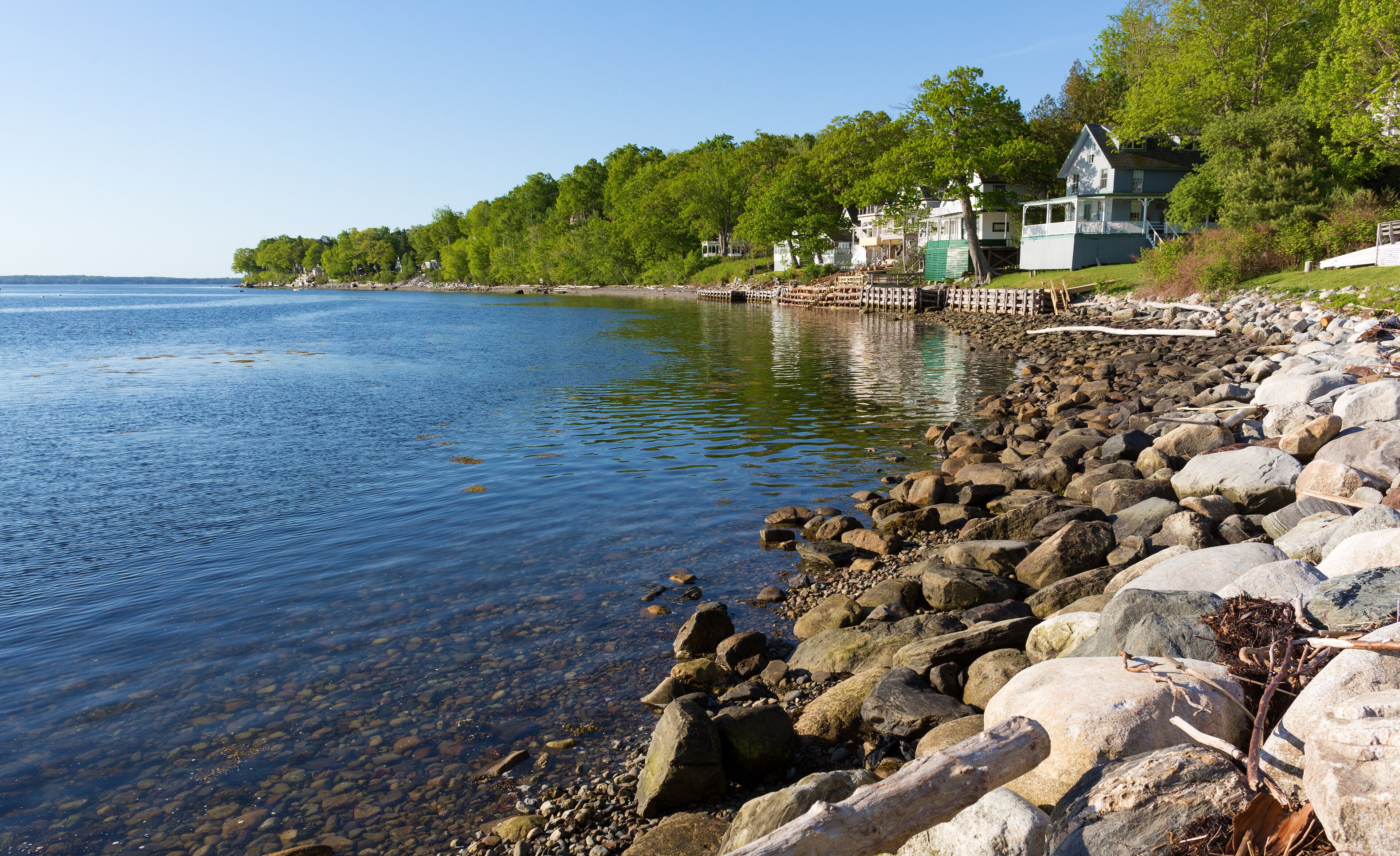 A rocky shoreline with clear water, a wooden dock, and houses in the background.