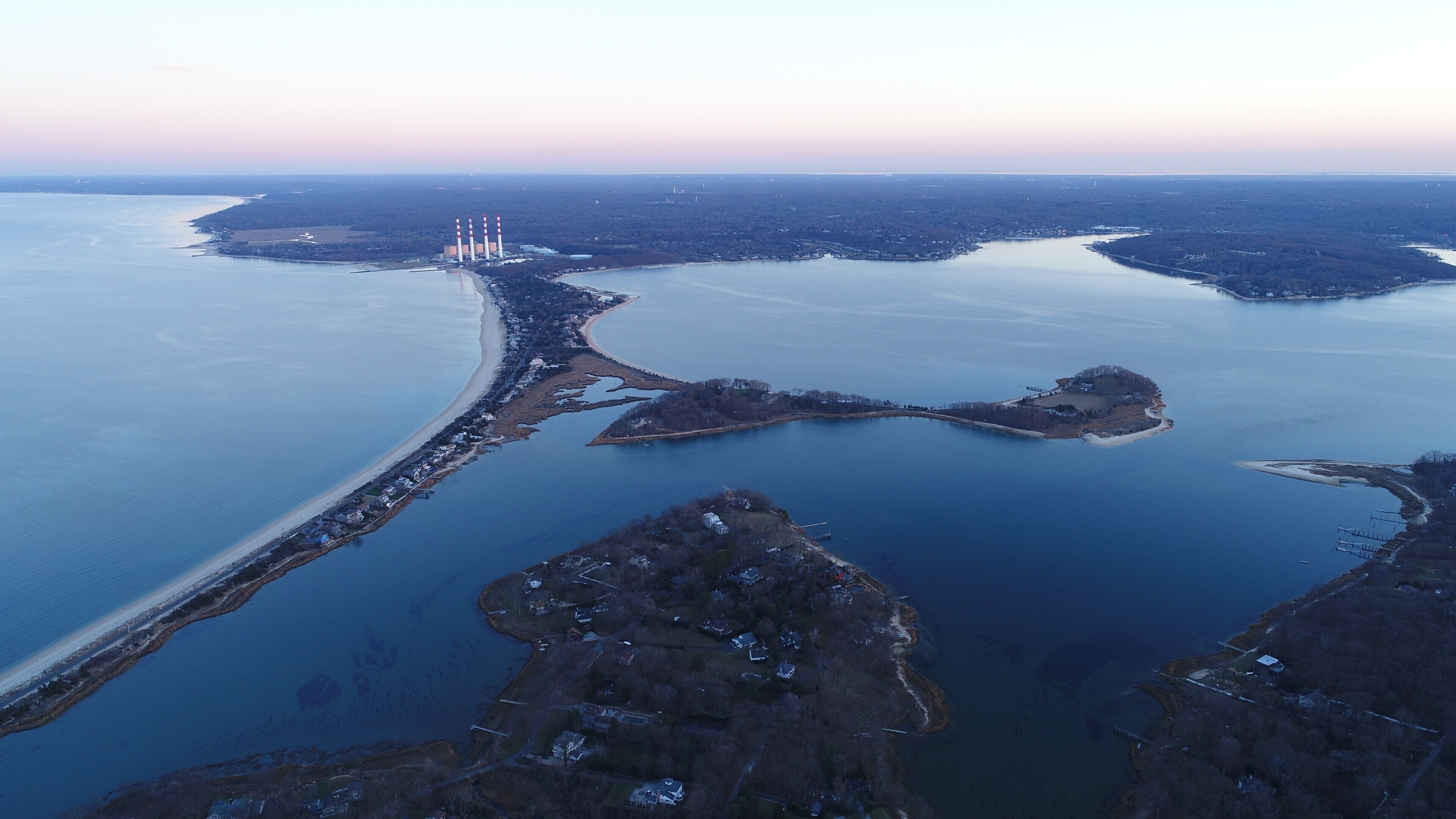 An aerial view of a coastal area with a large body of water, a sandy beach, and a series of industrial chimneys.