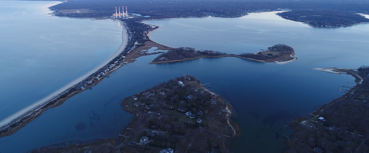 An aerial view of a coastal area with a large body of water, a sandy beach, and a series of industrial chimneys.