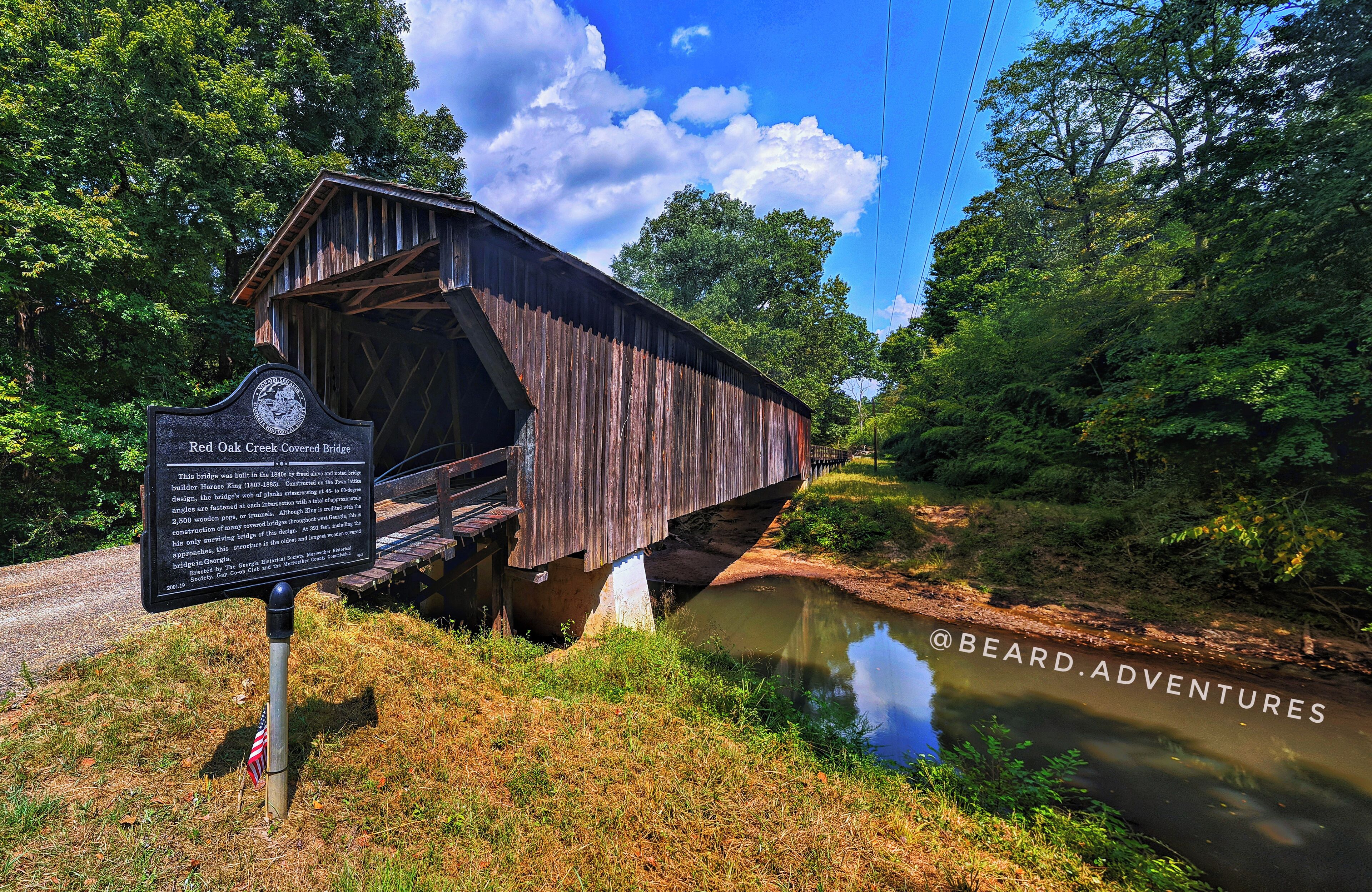 A covered wooden bridge over a river, surrounded by lush greenery and a clear blue sky.