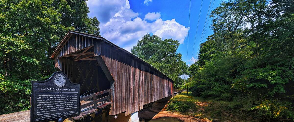 A covered wooden bridge over a river, surrounded by lush greenery and a clear blue sky.