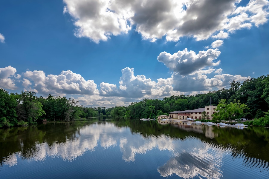 Un lac paisible où se reflètent les nuages, entouré d’une forêt dense et verdoyante, et un bâtiment coiffé d’un toit rouge.
