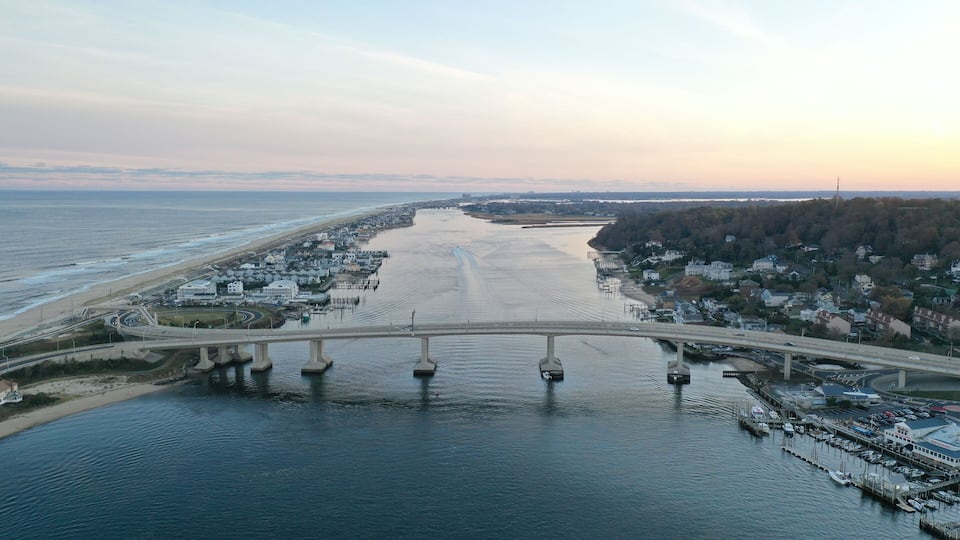 A coastal bridge over a wide river, connecting a beach and a town with buildings and docks.