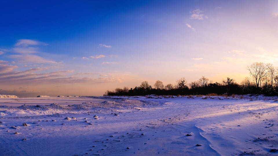 A snowy landscape with a frozen body of water, trees, and a clear sky.