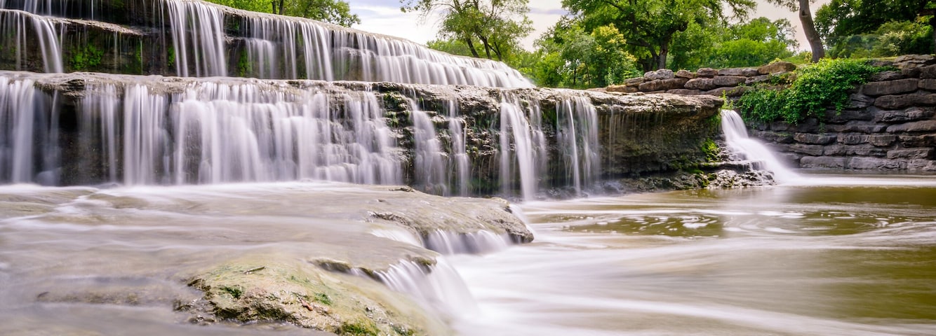 A waterfall cascading over rocks, surrounded by trees and a clear sky.