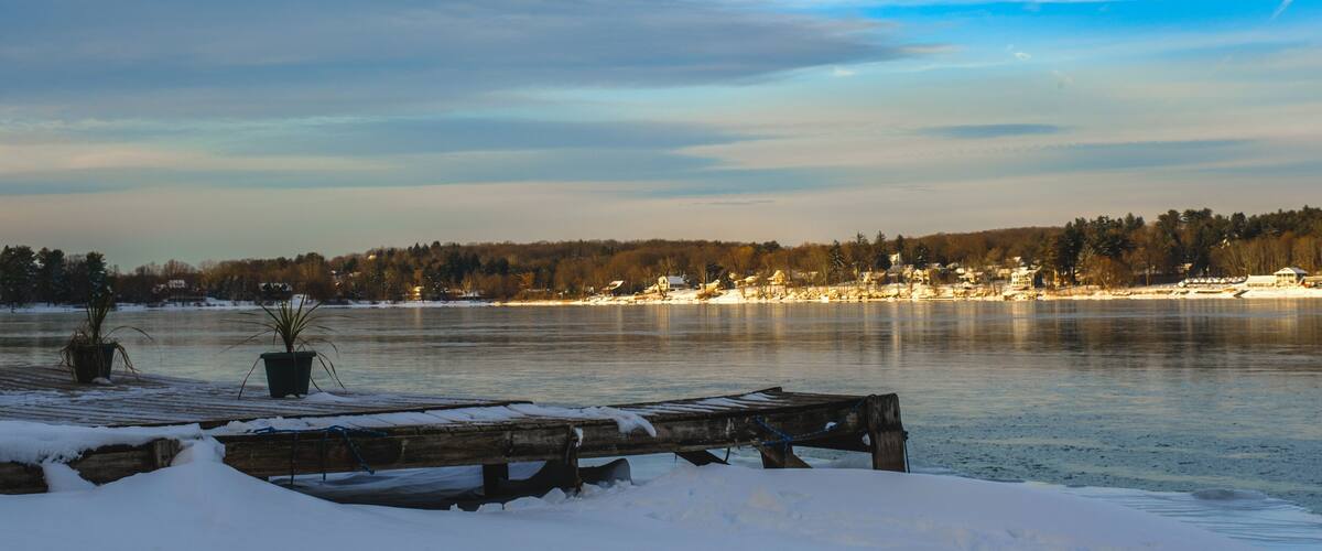 A frozen lake with a wooden dock and potted plants in the foreground, surrounded by trees and buildings in the background.