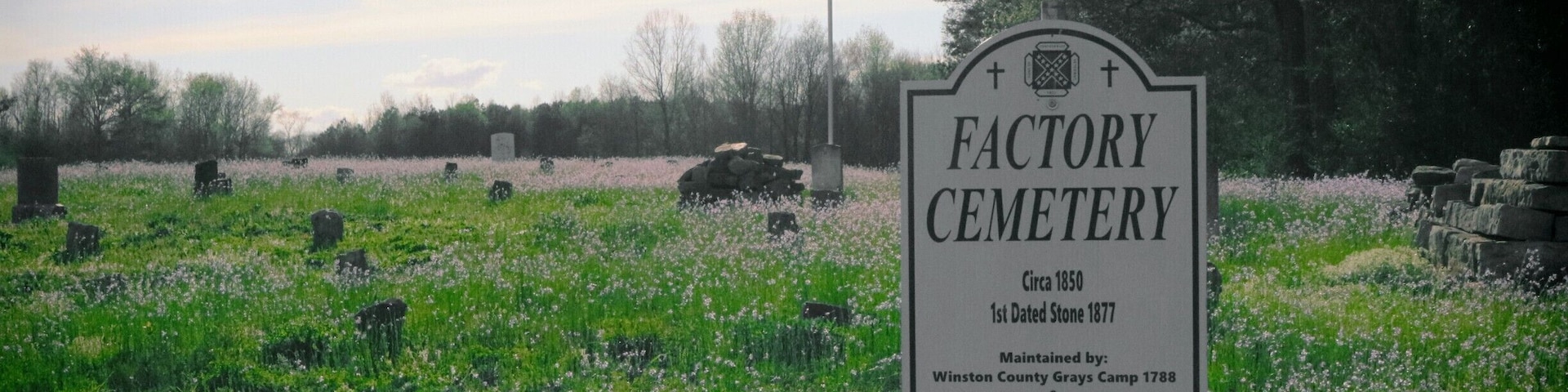 A cemetery sign with a flag, surrounded by gravestones and blooming flowers.