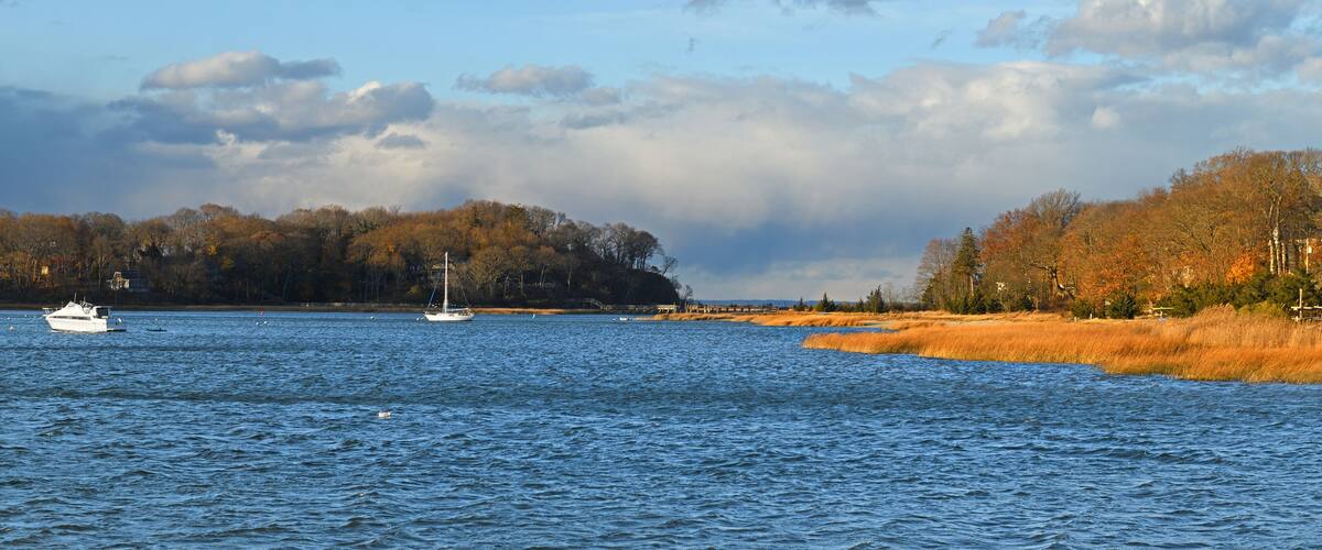 A calm lake with a sailboat, surrounded by autumn-colored trees and a cloudy sky.