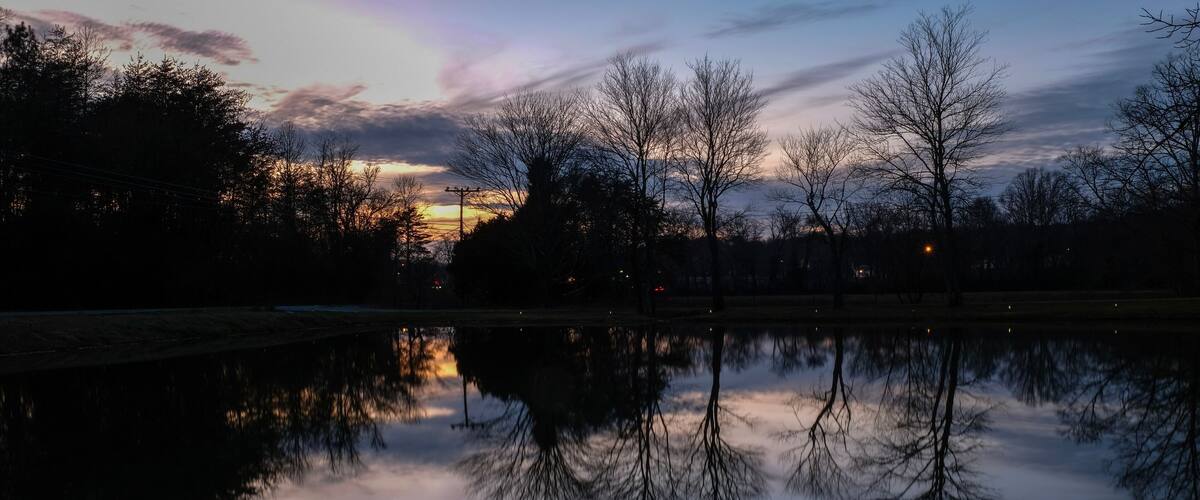 A calm lake at dusk with trees reflecting in the water and a sunset in the background.