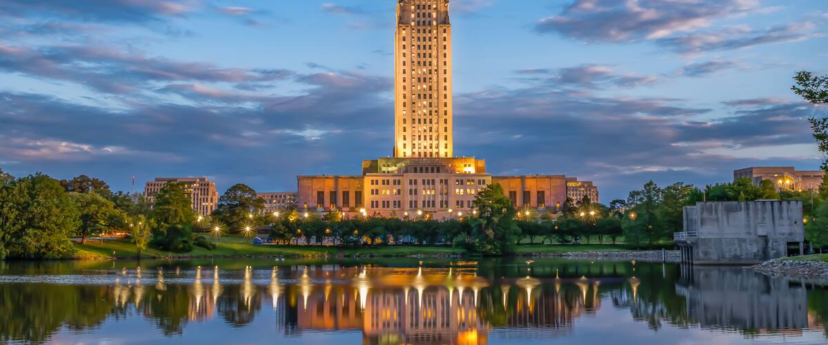 A tall, illuminated building with a spire, reflecting in a calm body of water, surrounded by greenery and other buildings.