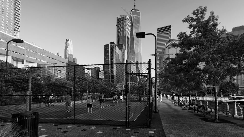 A city park with a basketball court, surrounded by tall buildings, including a distinctive skyscraper.