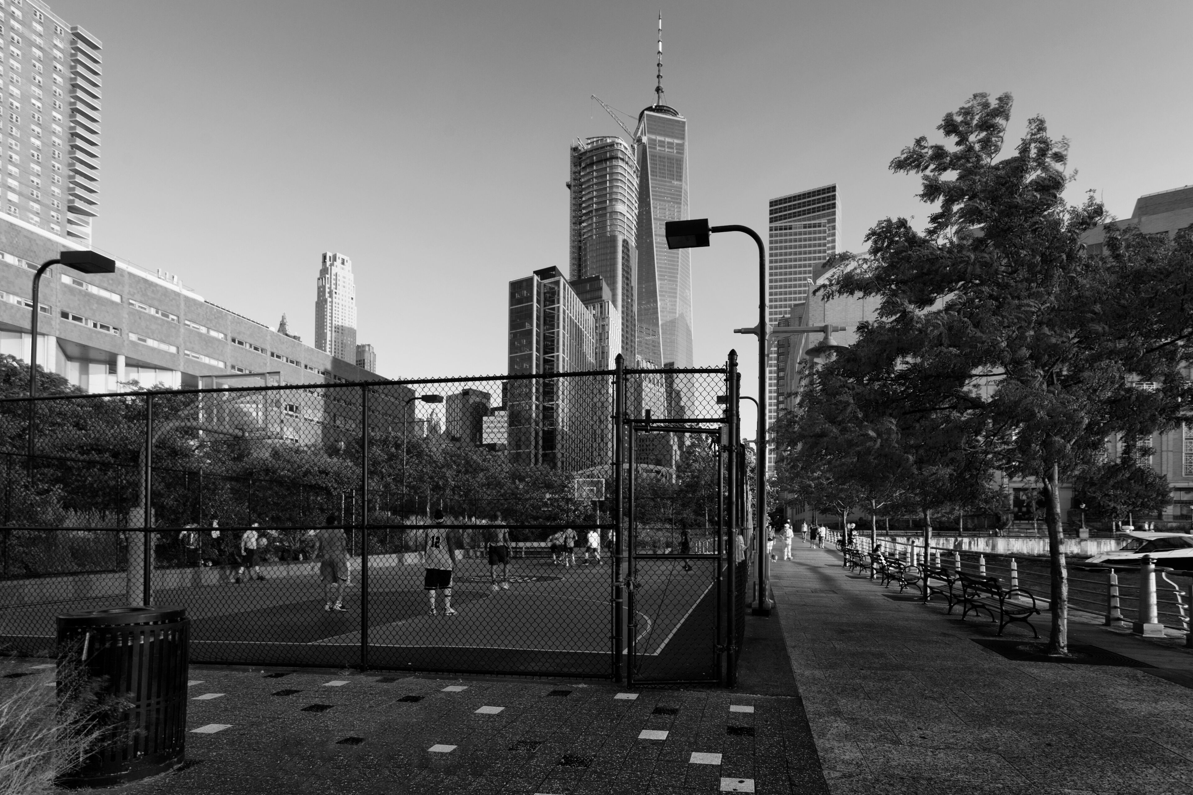 A city park with a basketball court, surrounded by tall buildings, including a distinctive skyscraper.