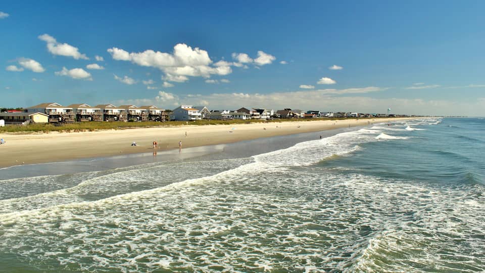A beach with waves, a sandy shore, and houses in the background.