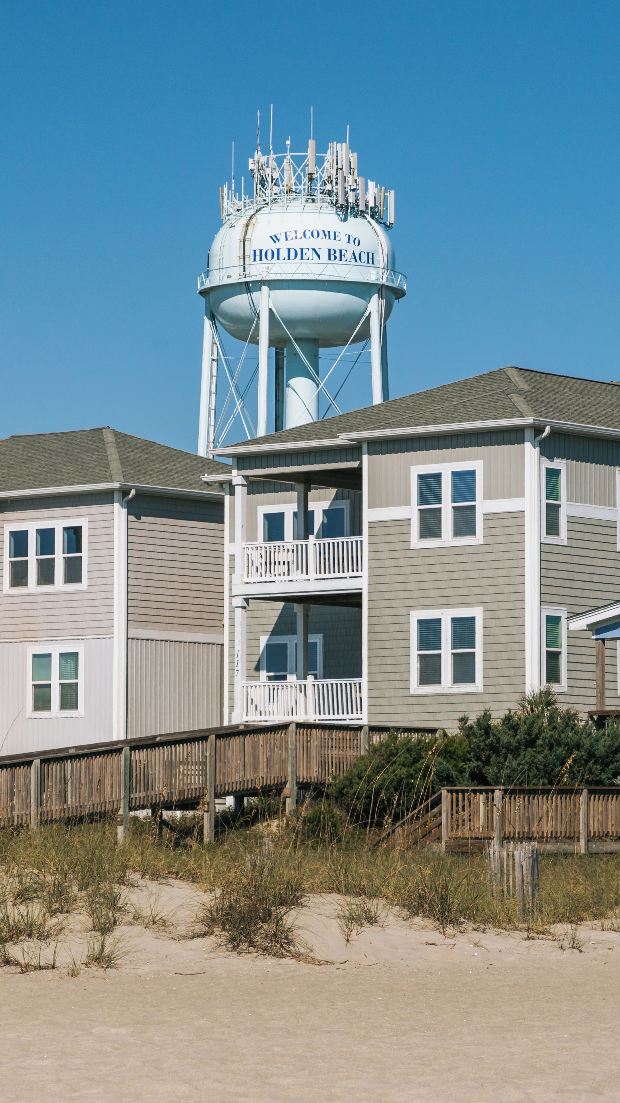 A water tower with 'Welcome to Holden Beach' written on it, a beachfront house, and a wooden boardwalk.