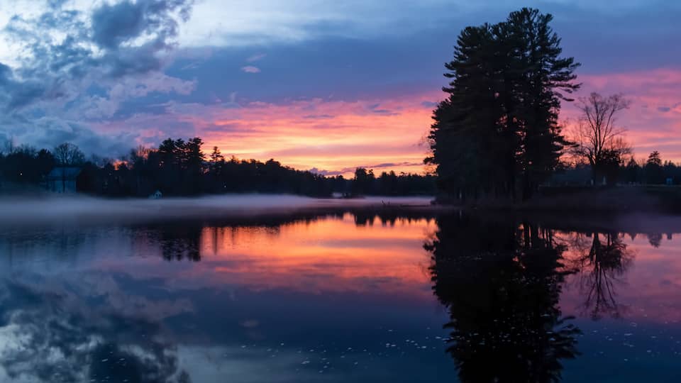 A calm lake at sunset with a reflection of trees and a clear sky.