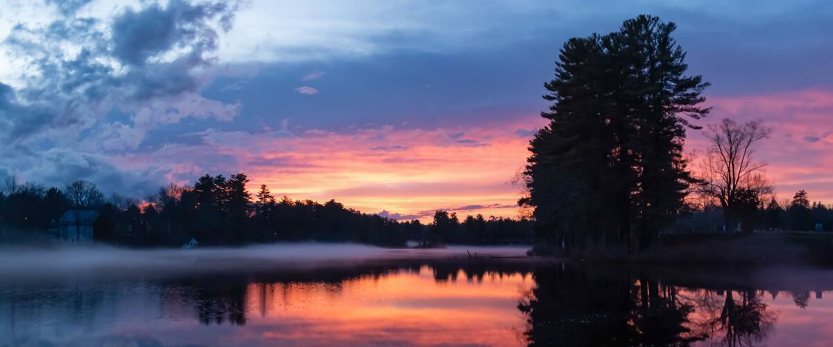 A calm lake at sunset with a reflection of trees and a clear sky.