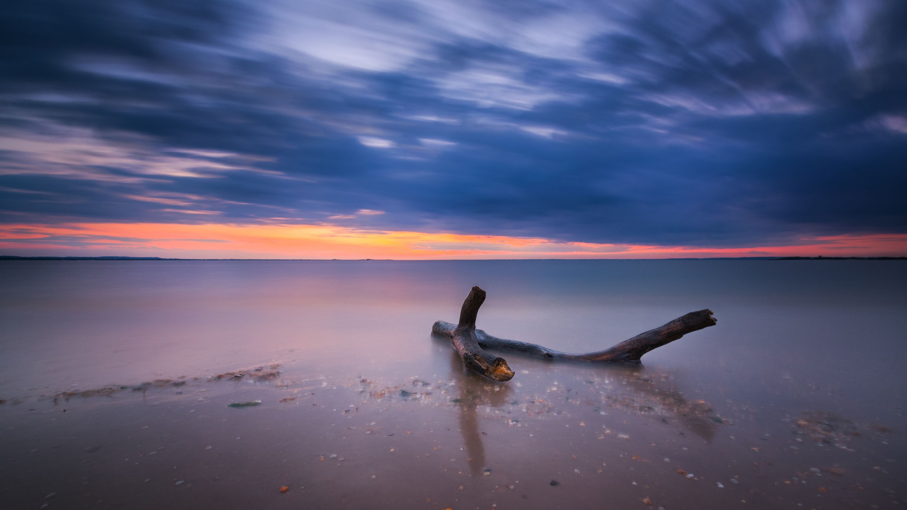 A calm body of water with a fallen tree trunk in the foreground and a colorful sunset in the background.