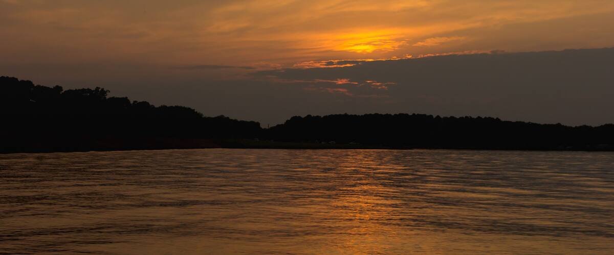 A sunset over a calm body of water with a silhouette of a forested hill in the background.
