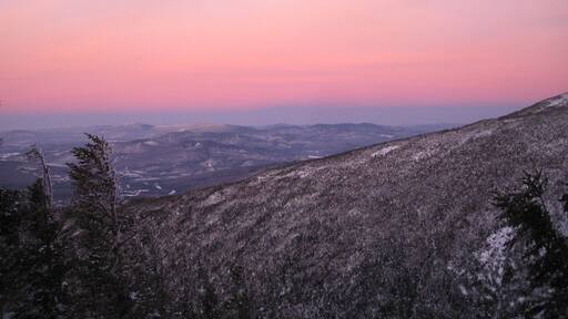 Pendiente nevada de montaña con vistas a valles lejanos y un cielo rosado al atardecer.
