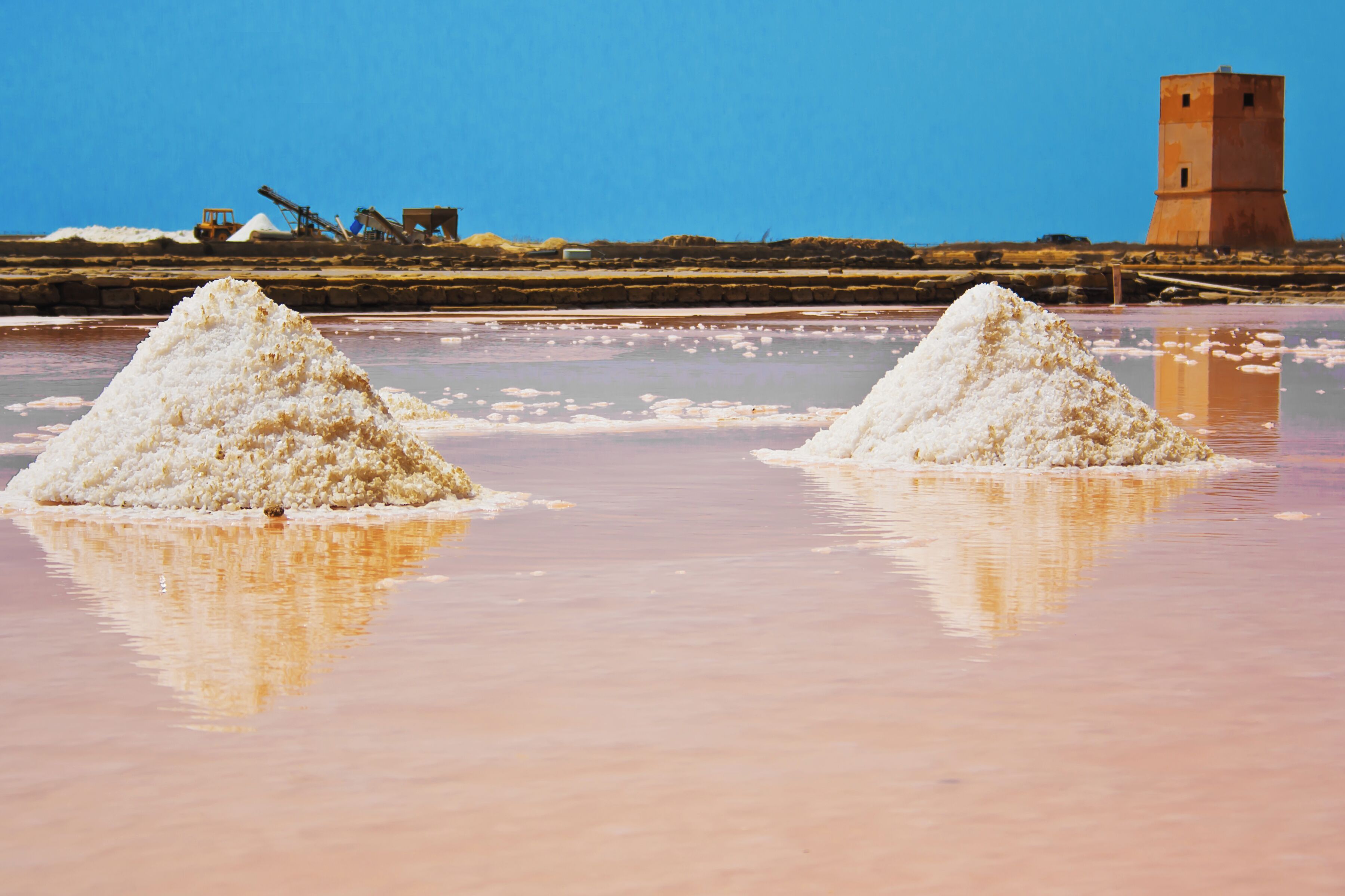 Two salt piles in a shallow body of water with a clear sky and a distant tower.