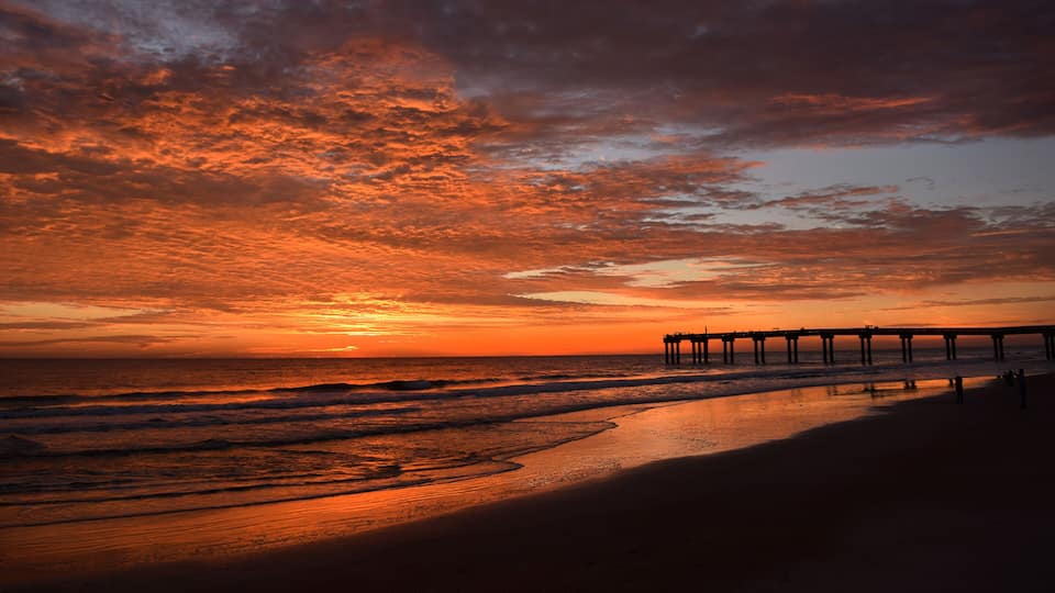 A sunset over a beach with waves, a pier extending into the water, and a clear sky.