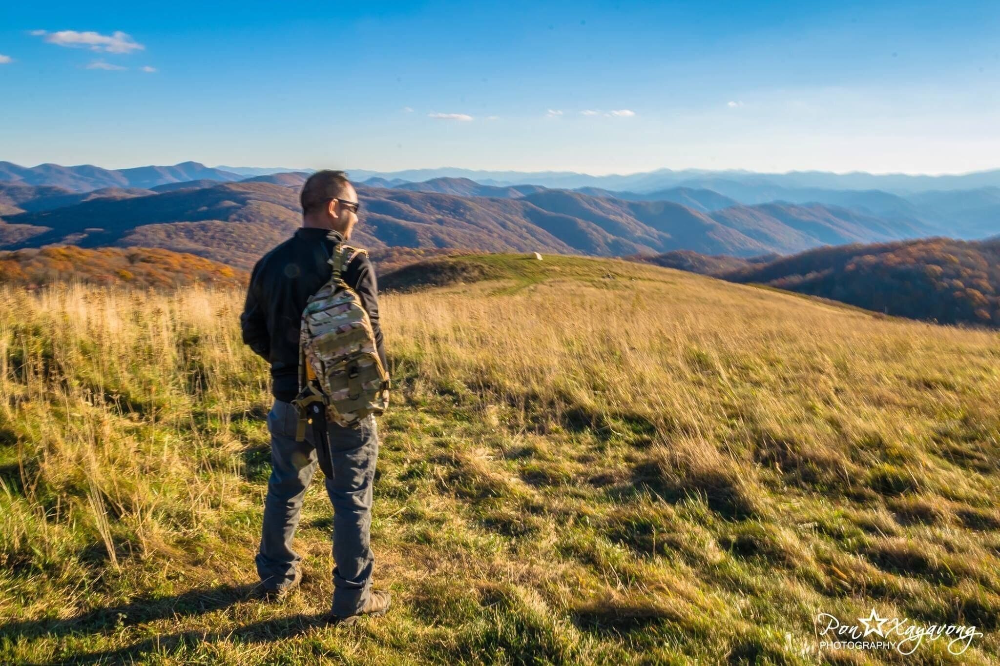 A person standing on a grassy hill with a mountainous landscape in the background.