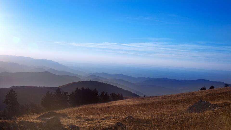 A mountainous landscape with rolling hills and sparse trees under a clear blue sky.