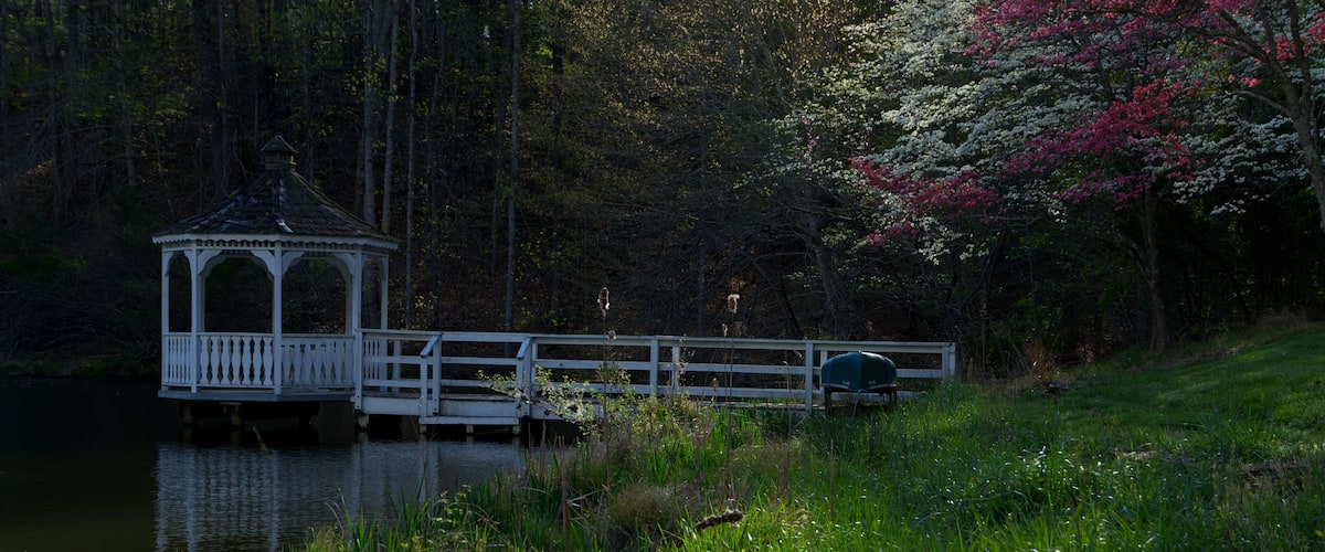 a tranquil pond on a calm spring morning with dogwood trees and red bud trees in full bloom with a canoe, a dock and a gazebo in the Blue Ridge Mountains of Virginia