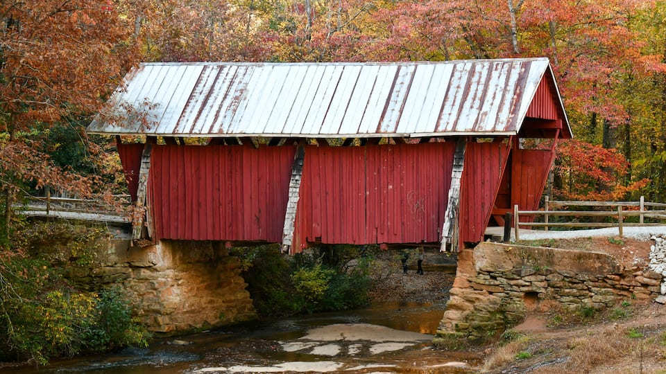 A red covered bridge over a small stream in a forested area with autumn foliage.