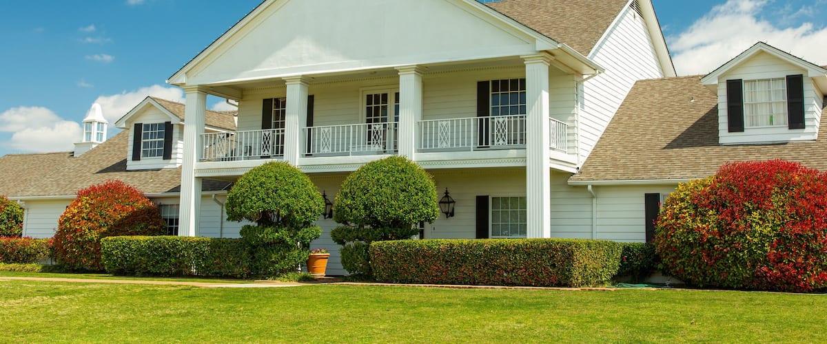 A large white house with a porch, two trees, and red bushes.