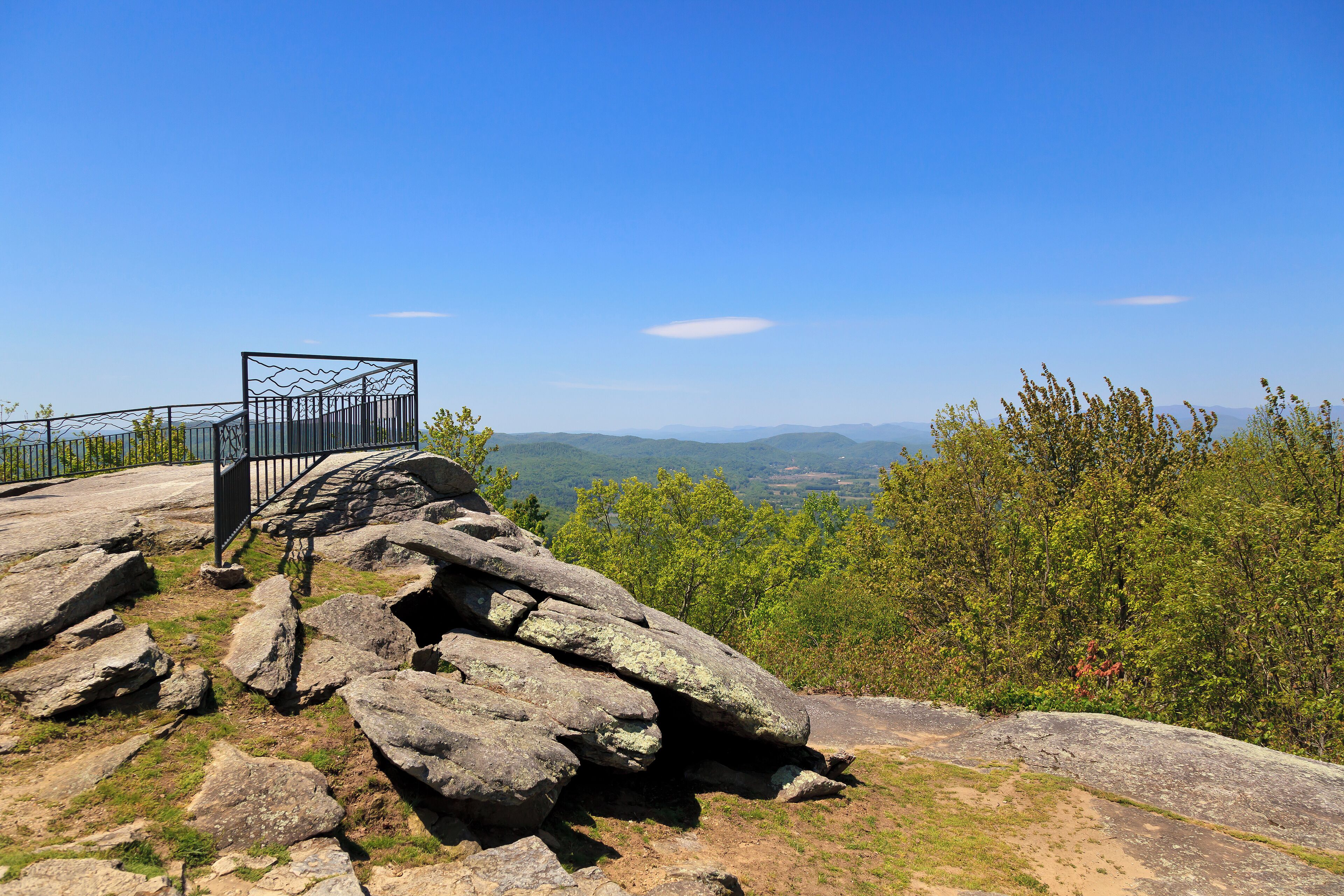 A rocky overlook with a metal railing, surrounded by trees and a clear blue sky.