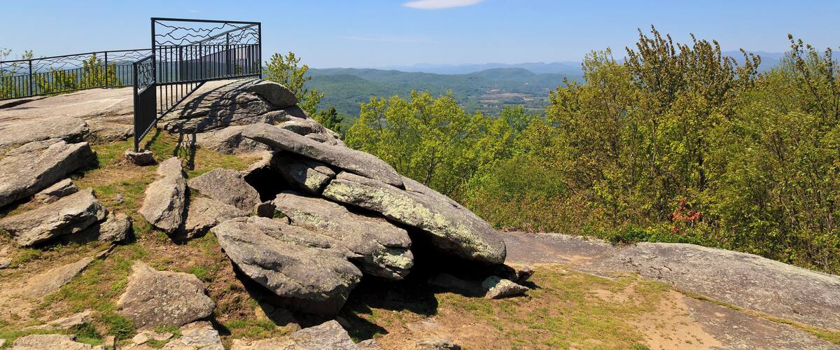 A rocky overlook with a metal railing, surrounded by trees and a clear blue sky.