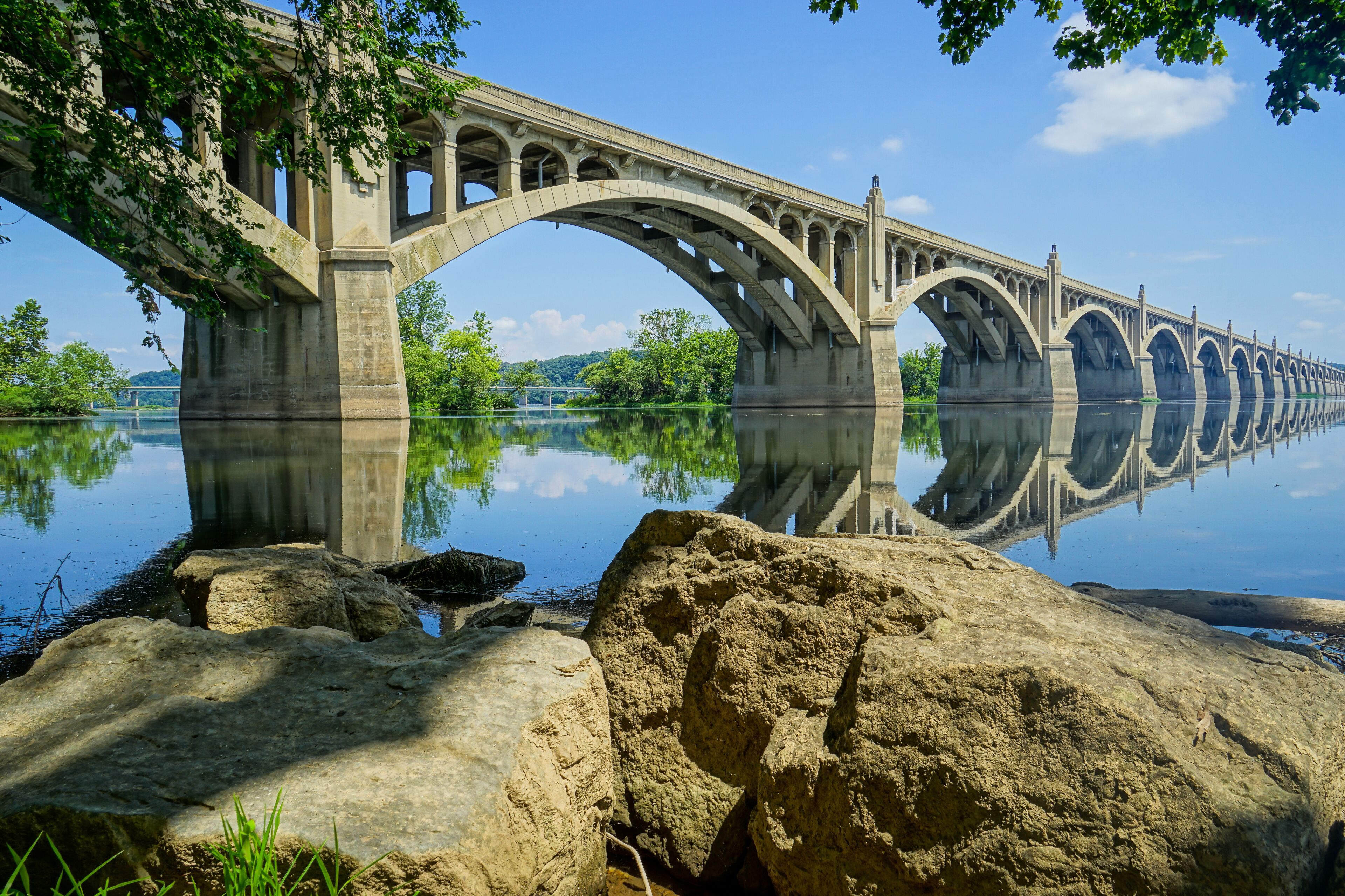 A large stone bridge spanning a calm river, with large rocks in the foreground and trees on the banks.