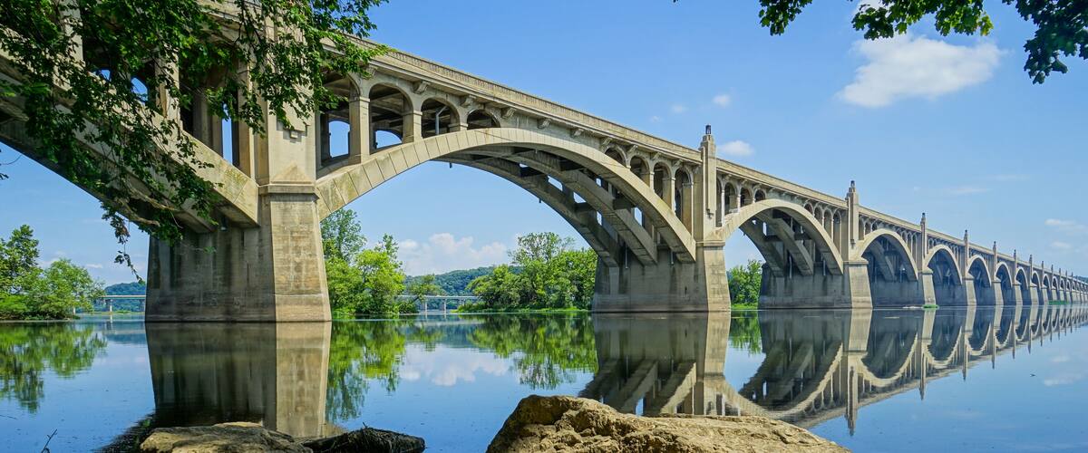 A large stone bridge spanning a calm river, with large rocks in the foreground and trees on the banks.
