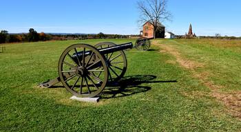 A cannon in a grassy field with a wooden house and a monument in the background.