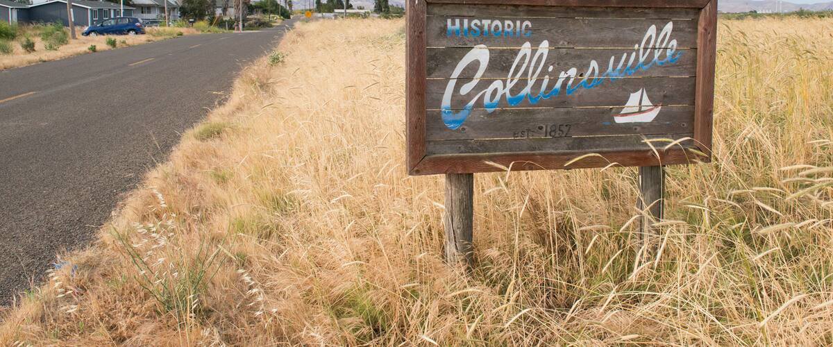 A wooden sign with 'Historic Collinsville' written on it, surrounded by dry grass and a road with houses in the background.