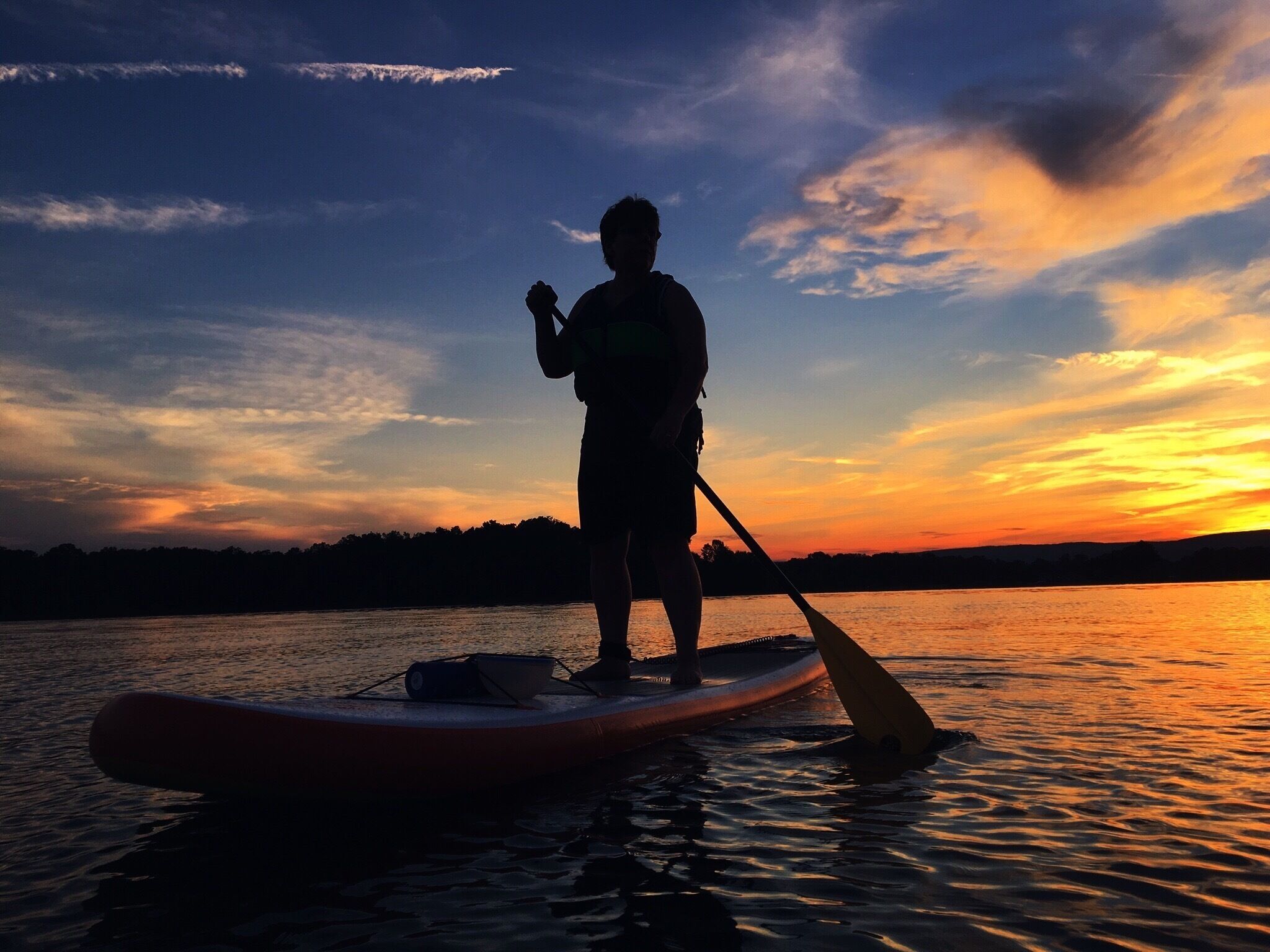 A person paddleboarding at sunset on a calm body of water with a silhouette of trees in the background.
