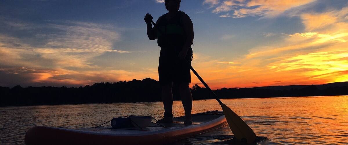 A person paddleboarding at sunset on a calm body of water with a silhouette of trees in the background.