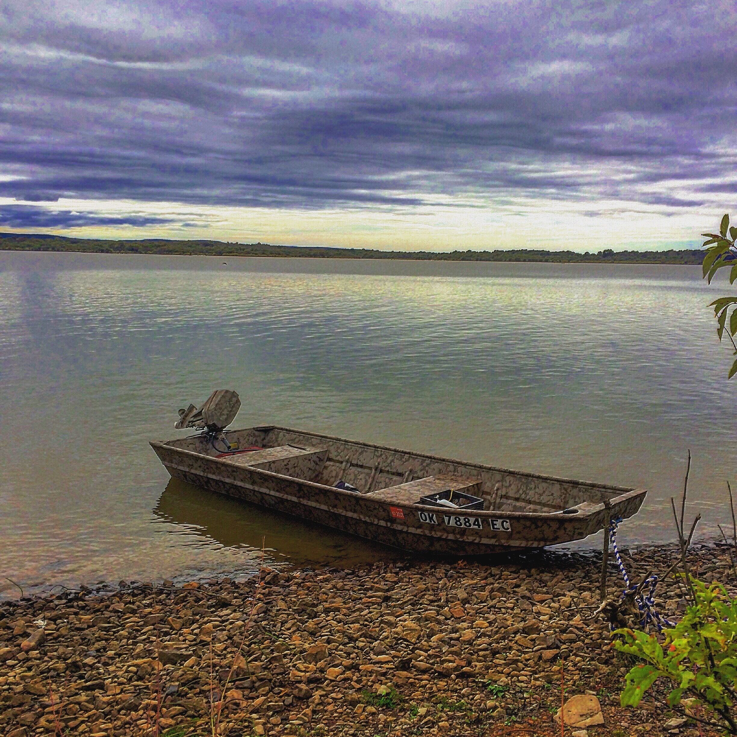 A small boat on a calm lake with a rocky shore and a cloudy sky.
