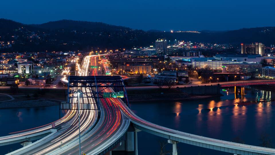 A nighttime cityscape with a multi-level highway and a river reflecting city lights.