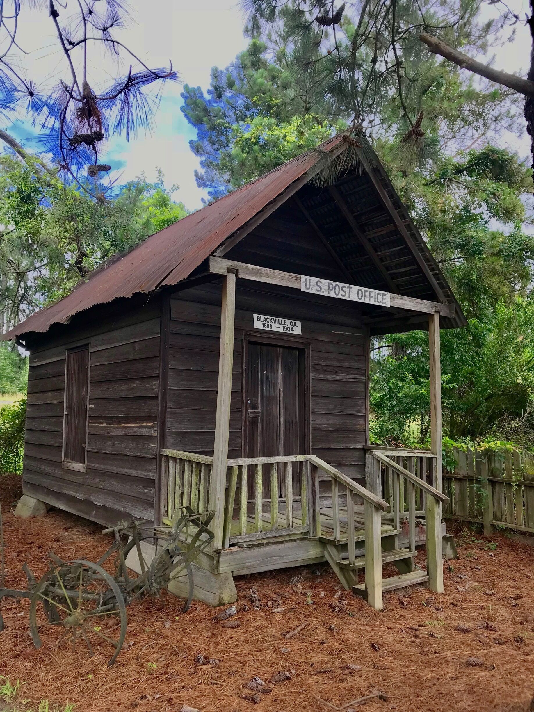 A rustic U.S. Post Office building in Blackville, GA., surrounded by pine trees and a wooden fence.