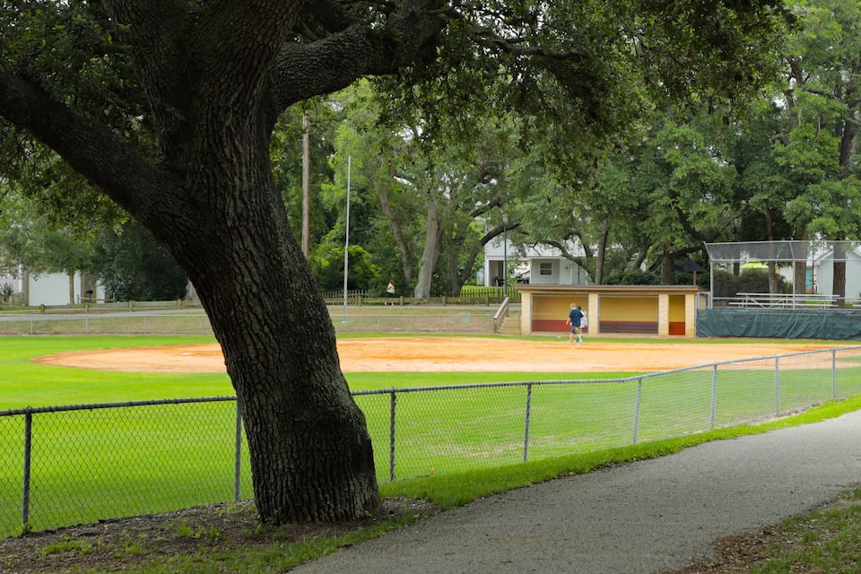 A baseball field with a large tree in the foreground, a covered dugout, and a walking path along the edge.