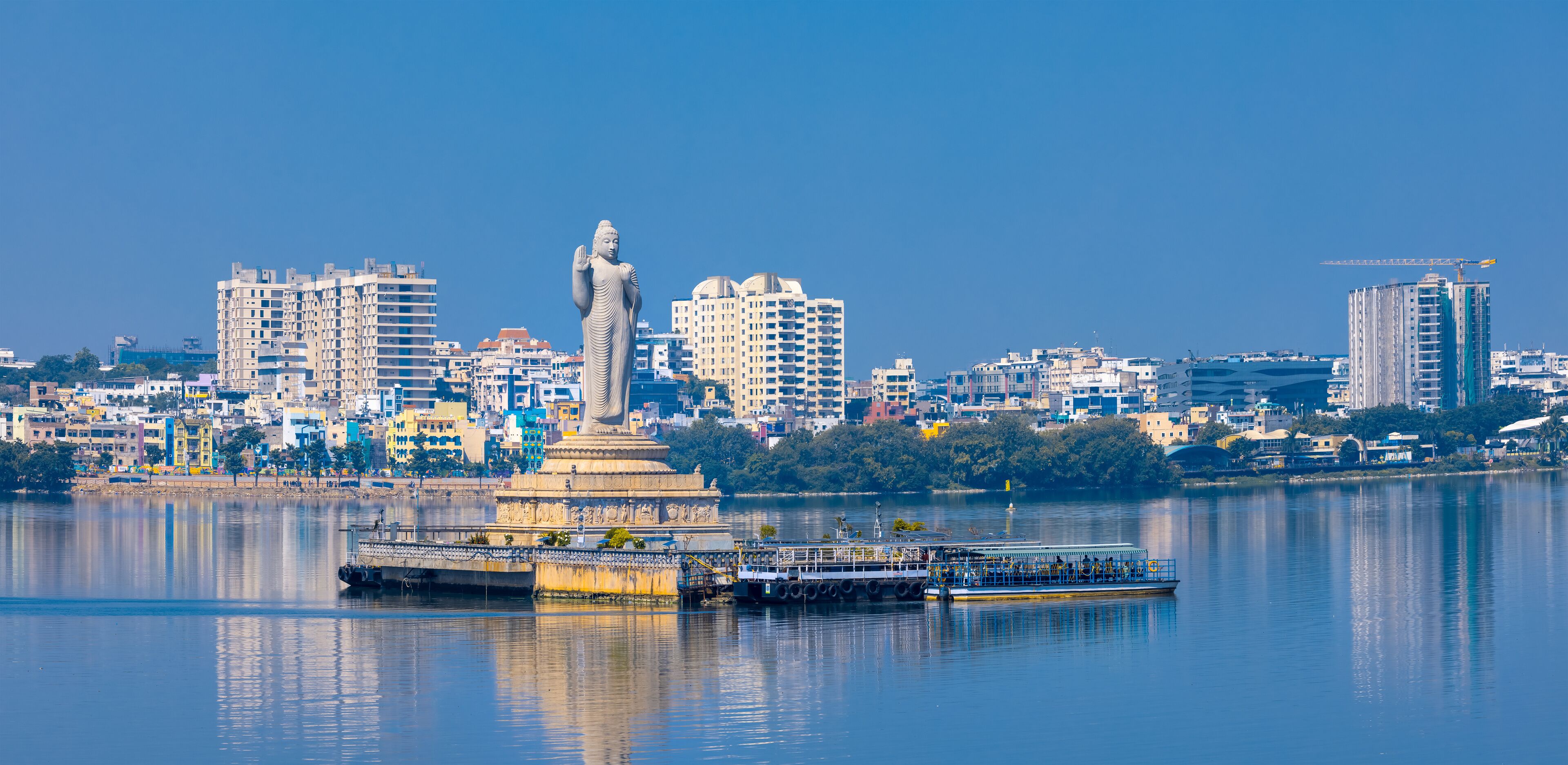 A statue in a city with a river, buildings, and a boat.