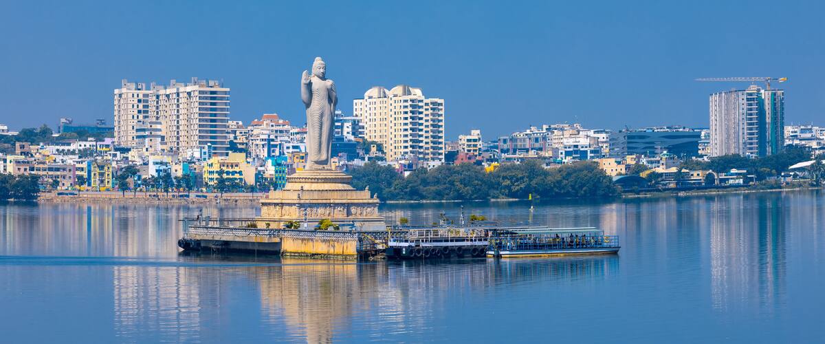 A statue in a city with a river, buildings, and a boat.