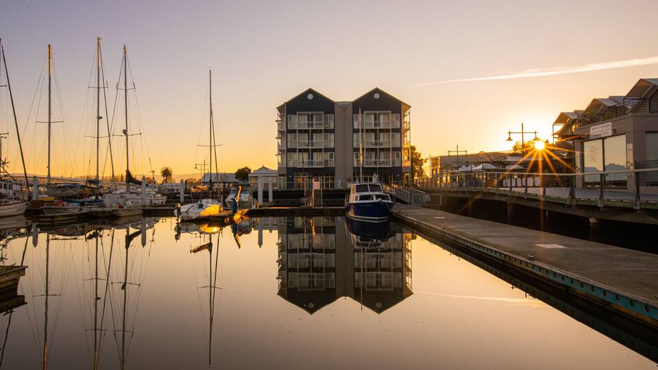 A marina with boats docked, a bridge, and buildings reflecting in the water during sunset.
