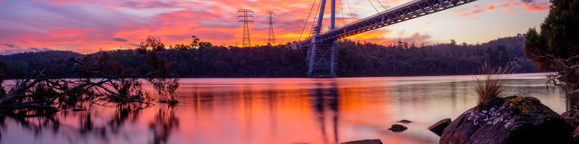 A suspension bridge over a calm lake at sunset with a forested shoreline.
